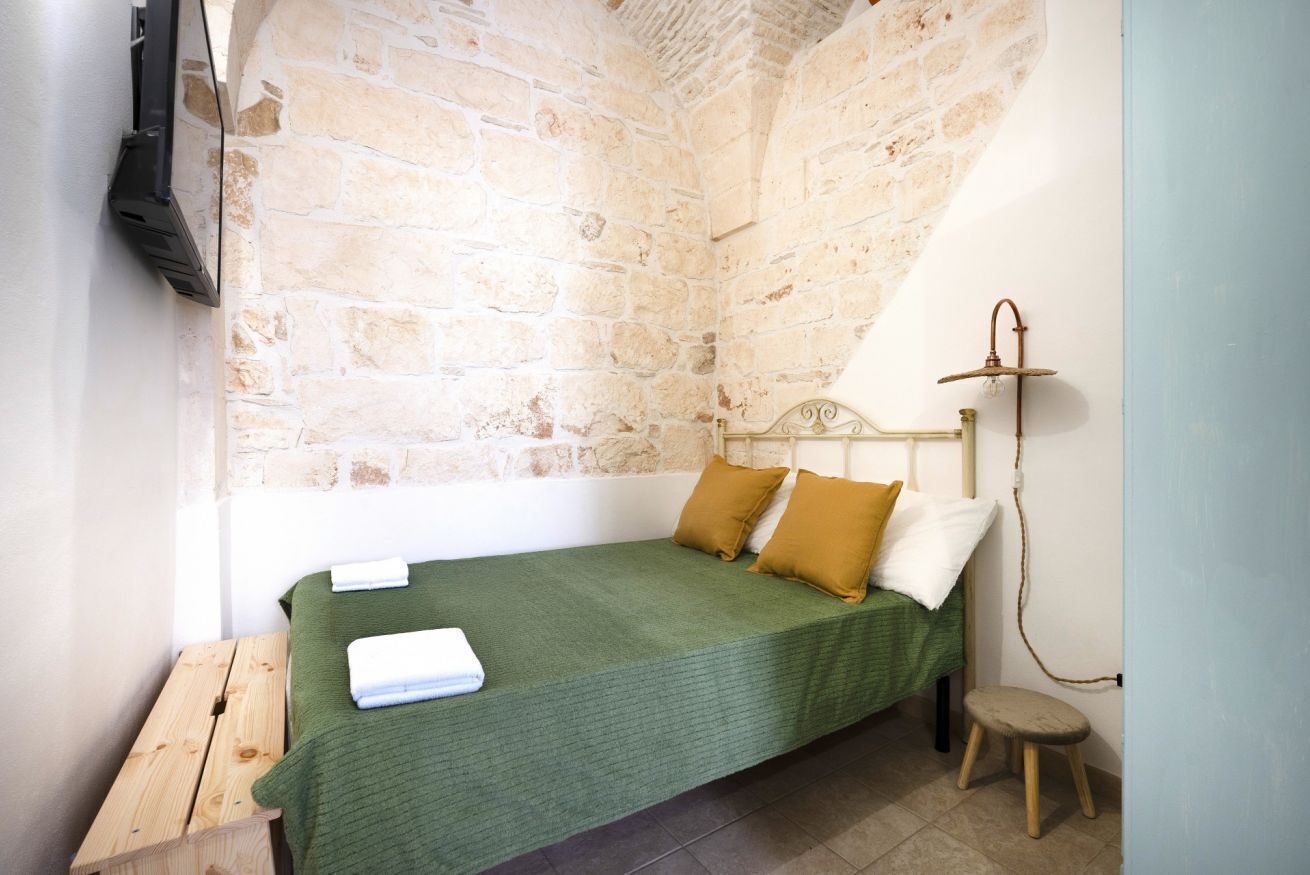 A small, cozy room with a stone wall, a bed with green bedding, a wooden bench, and a small stool.