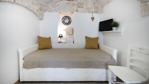 Une chambre avec un plafond en pierre, un lit recouvert d’une couverture beige, une horloge murale, un téléviseur et une petite étagère avec une plante.