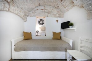 A bedroom with a stone ceiling, a bed with a beige blanket, a wall clock, a TV, and a small shelf with a plant. - Cozy apartment on two levels that accommodate up to 3 people and is equipped with all the main comforts for a pleasant stay. The location is excellent, in the center, with all the essential services at hand and a short distance from the points of interes (Martina Franca)