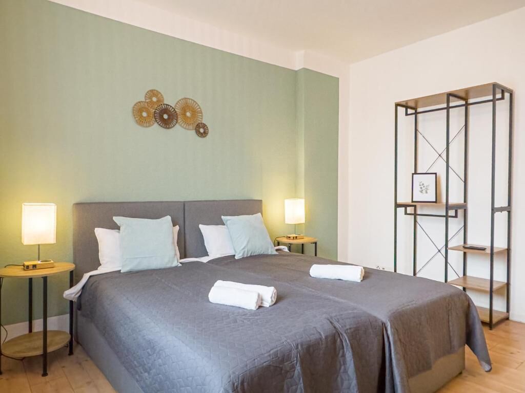 Una cama bien hecha con dos almohadas, dos mesitas de noche con lámparas, y un estante en la pared con un marco de fotos.