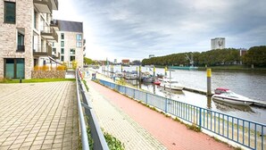 A riverside walkway with a paved path, a blue railing, and a row of boats docked along the water. - Marßeler Str. 21 (Bremen)
