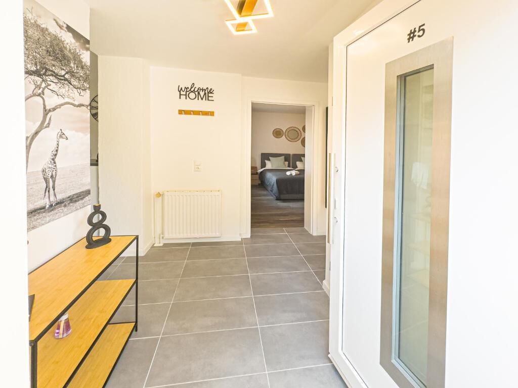 A hallway with a 'Welcome Home' sign, a number '5' on the wall, a radiator, and a bed in the background.