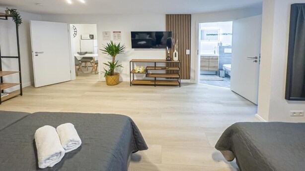 A hotel room with two beds, a TV, a plant, a wooden bench, and a view of the kitchen. - Marßeler Str. 21 (Bremen)