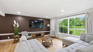 Ein modernes Wohnzimmer mit Flachbildfernseher, grauen Sofas, einem Couchtisch und einem großen Fenster mit Blick auf Grünflächen.