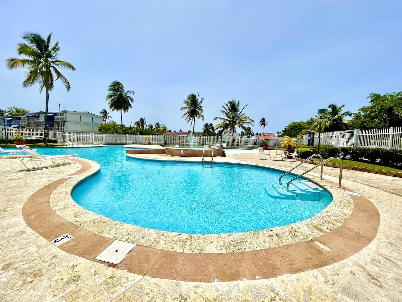 Una piscina de diseño curvo, rodeada de palmeras y una valla blanca.