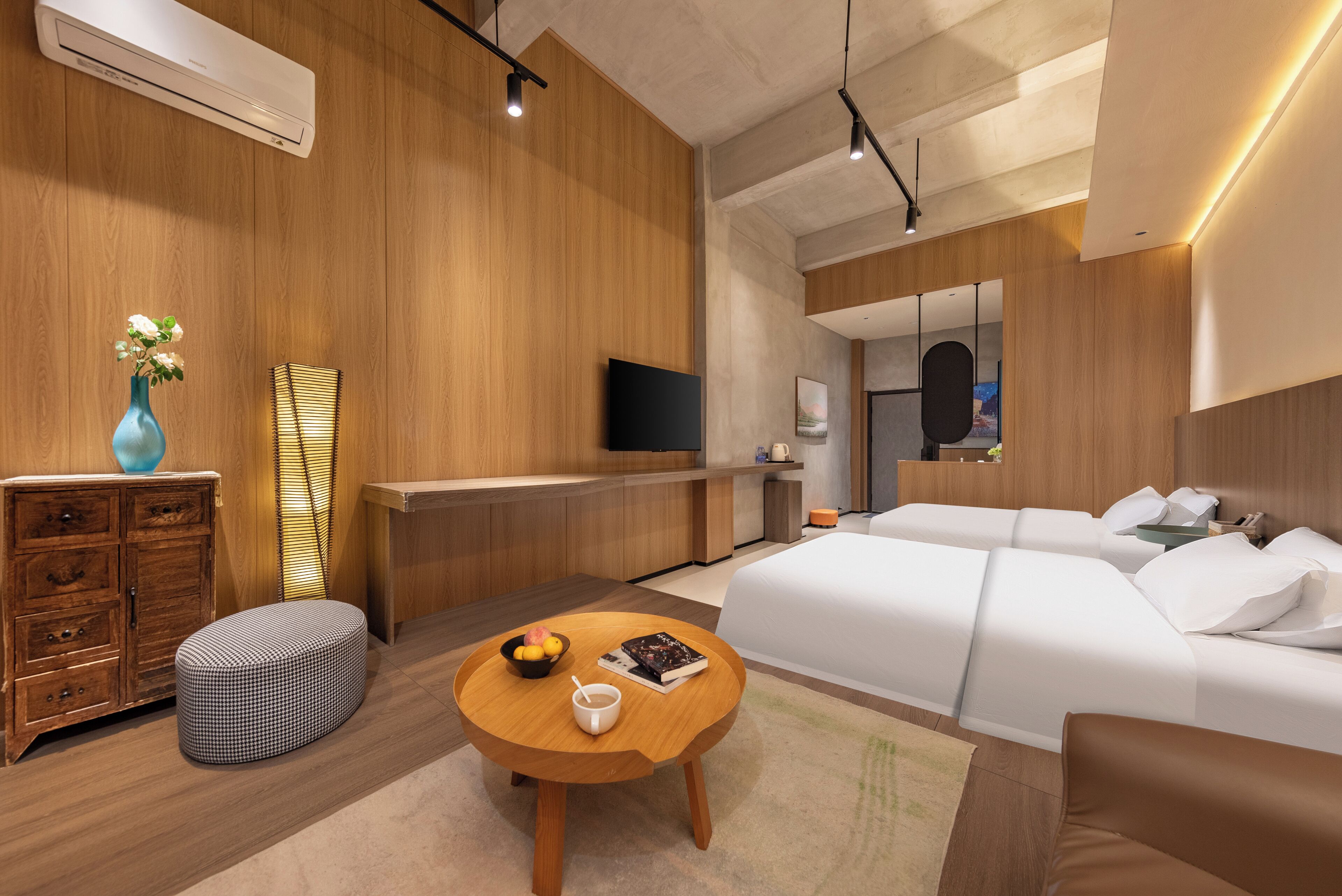A modern hotel room with a wooden wall, a flat-screen TV, a sofa, a dining table with a bowl of fruit and a book, and a bedside table with a lamp.
