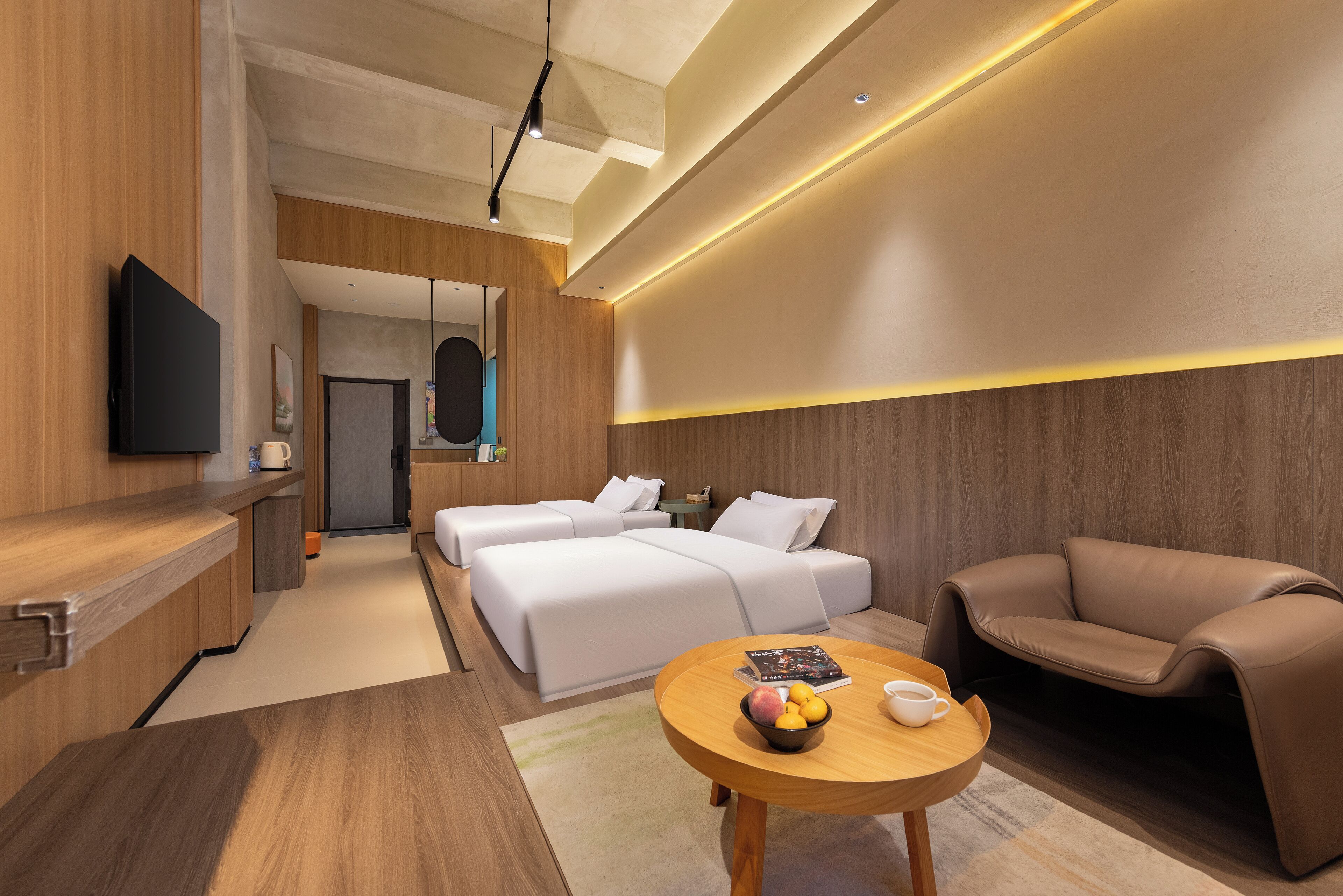 A modern hotel room with a wooden floor, a round wooden table with a bowl of fruit, a brown leather sofa, a bed with white linens, and a flat-screen TV mounted on the wall.