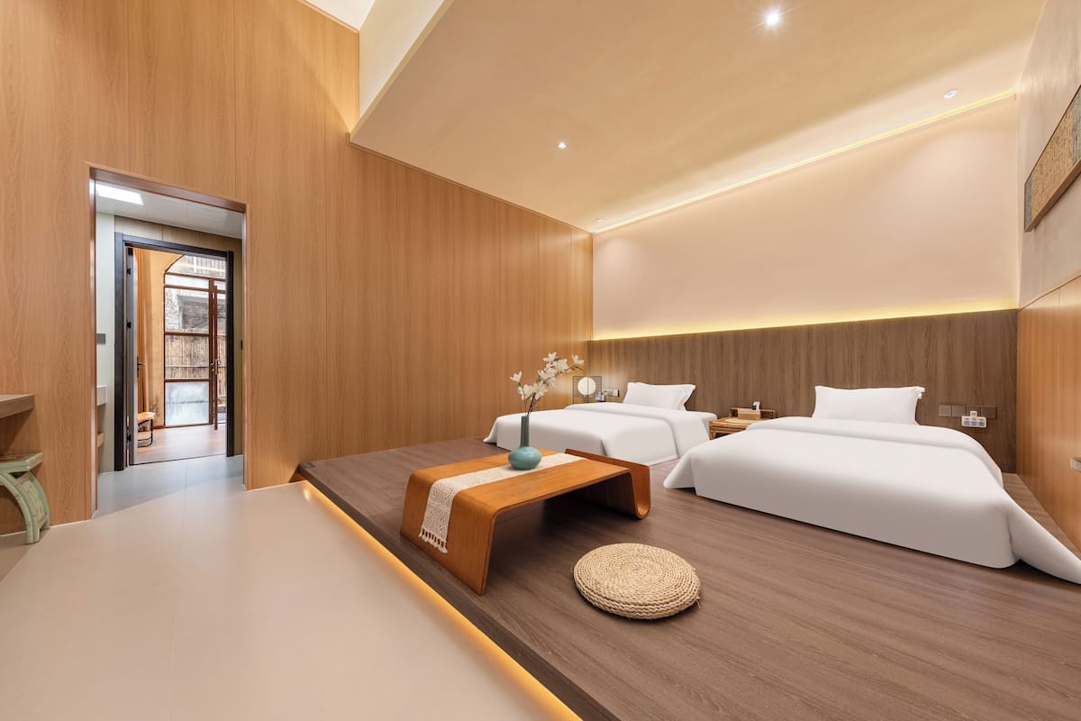 A modern hotel room with a wooden floor, two beds, a bench, a vase with flowers, and a view of a courtyard.