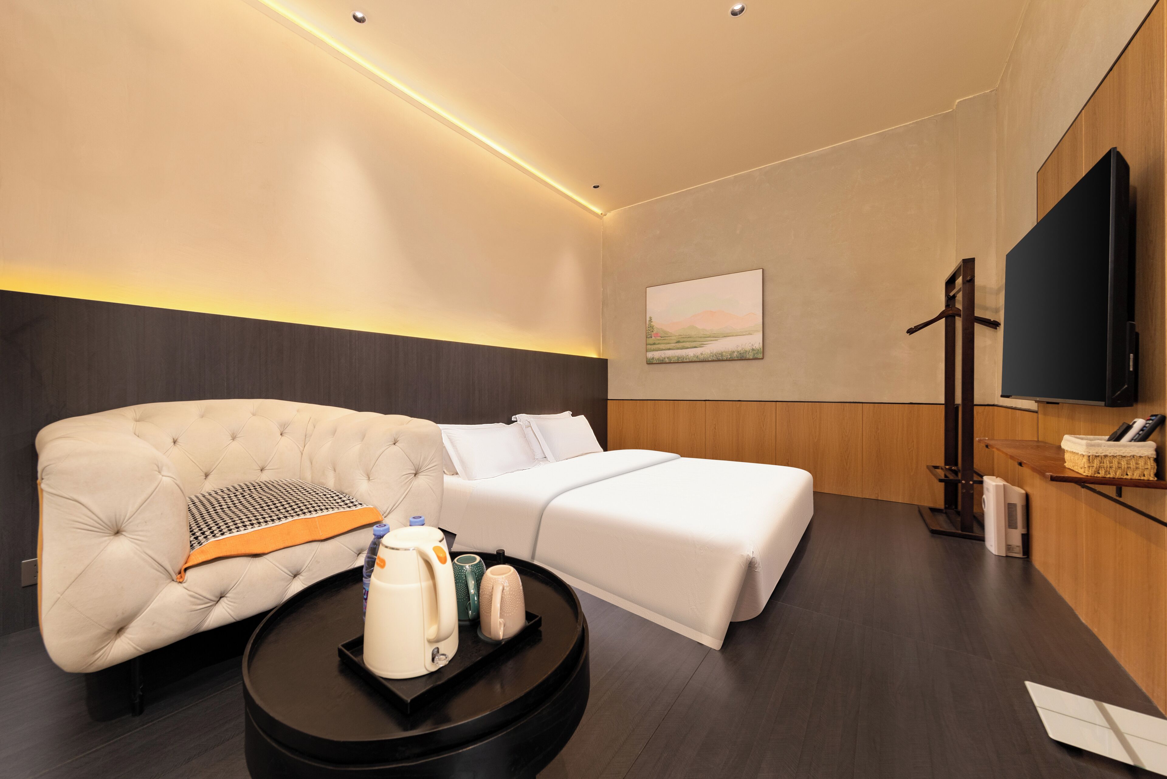 Una habitación de hotel moderna con sofá, cama, una mesita de centro con tetera y tazas, televisor de pantalla plana y un cuadro en la pared.