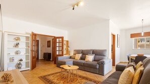 Een woonkamer met een grijze bank, een klein rond tafeltje, een kamerkleed met dessin en een houten kast met decoratieve objecten.