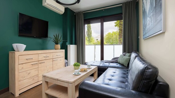 A modern living room with a wooden cabinet, a black leather sofa, a wooden coffee table, a flat-screen TV, and a large window with curtains. - Stylish Apartment with a Private Garden | Air Conditioning (Warszawa)