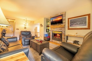 Ein gemütliches Wohnzimmer mit Ledersofas, einem Kamin, einem an einer Steinwand montierten Fernseher und einer Chandelüster.