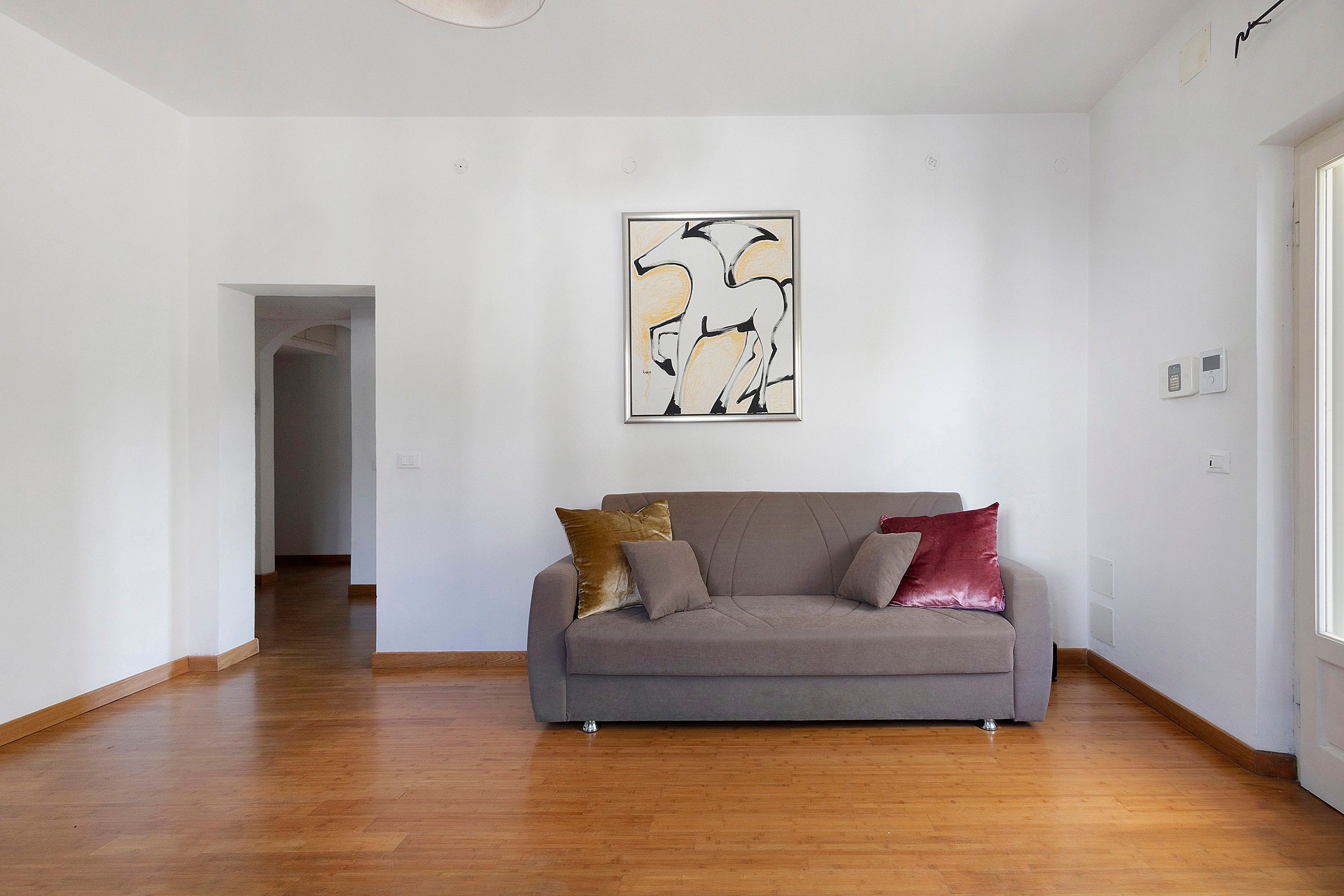 Una sala de estar con un sofá gris, dos cojines, piso de madera y una pared blanca con un cuadro abstracto enmarcado.
