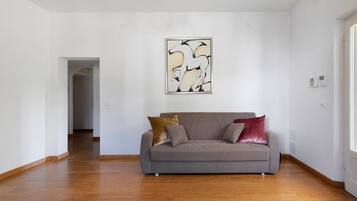 Una sala de estar con un sofá gris, dos cojines, piso de madera y una pared blanca con un cuadro abstracto enmarcado.