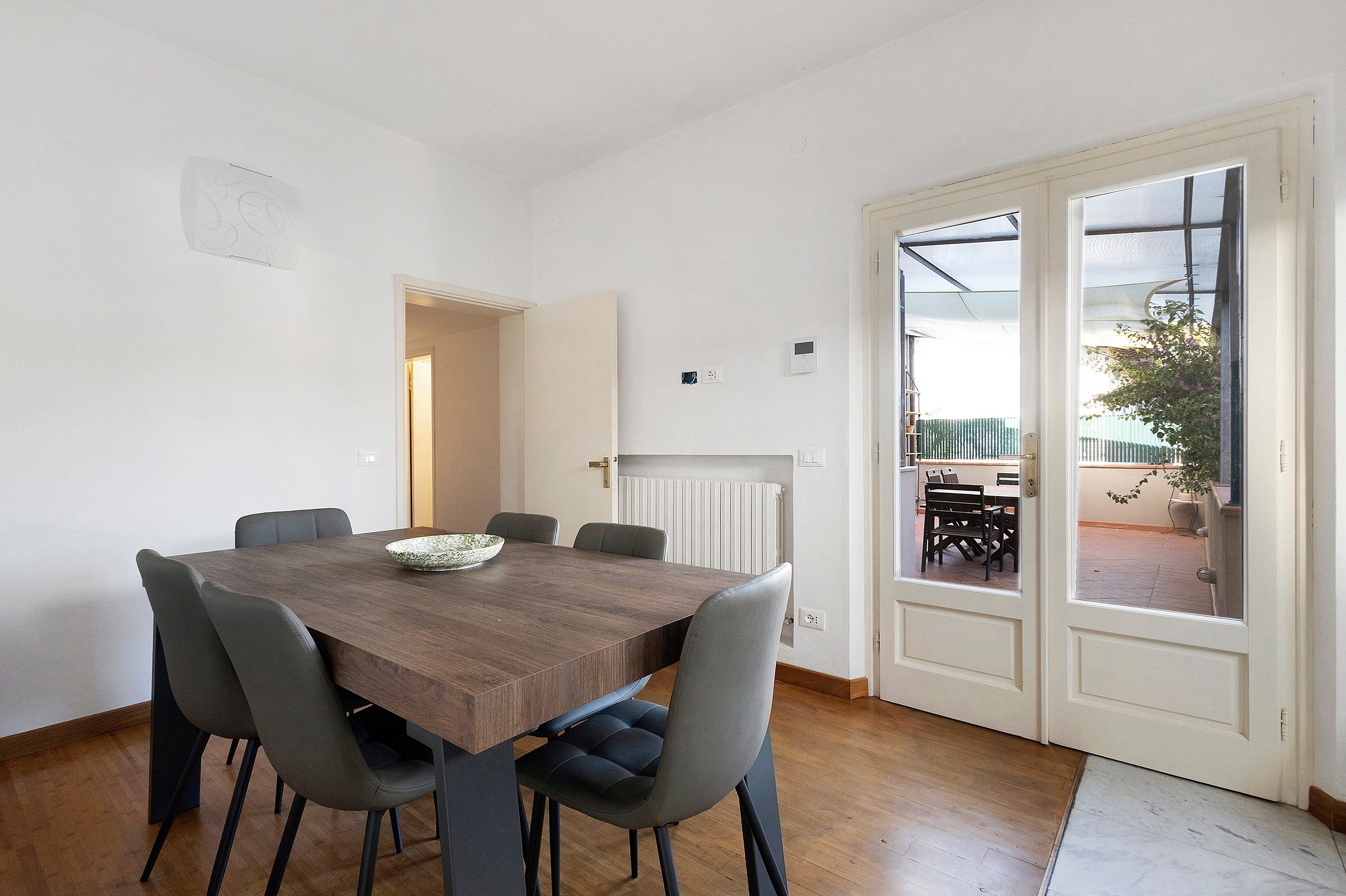 Een eetruimte met een houten tafel, zes stoelen en een schaal. Er is een deur naar buiten en een radiator aan de muur.