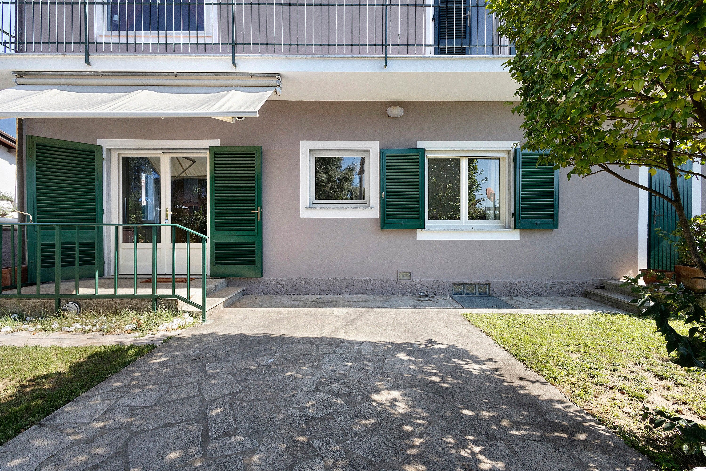 A two-story residential building with a covered entrance, green shutters, and a stone-paved front.