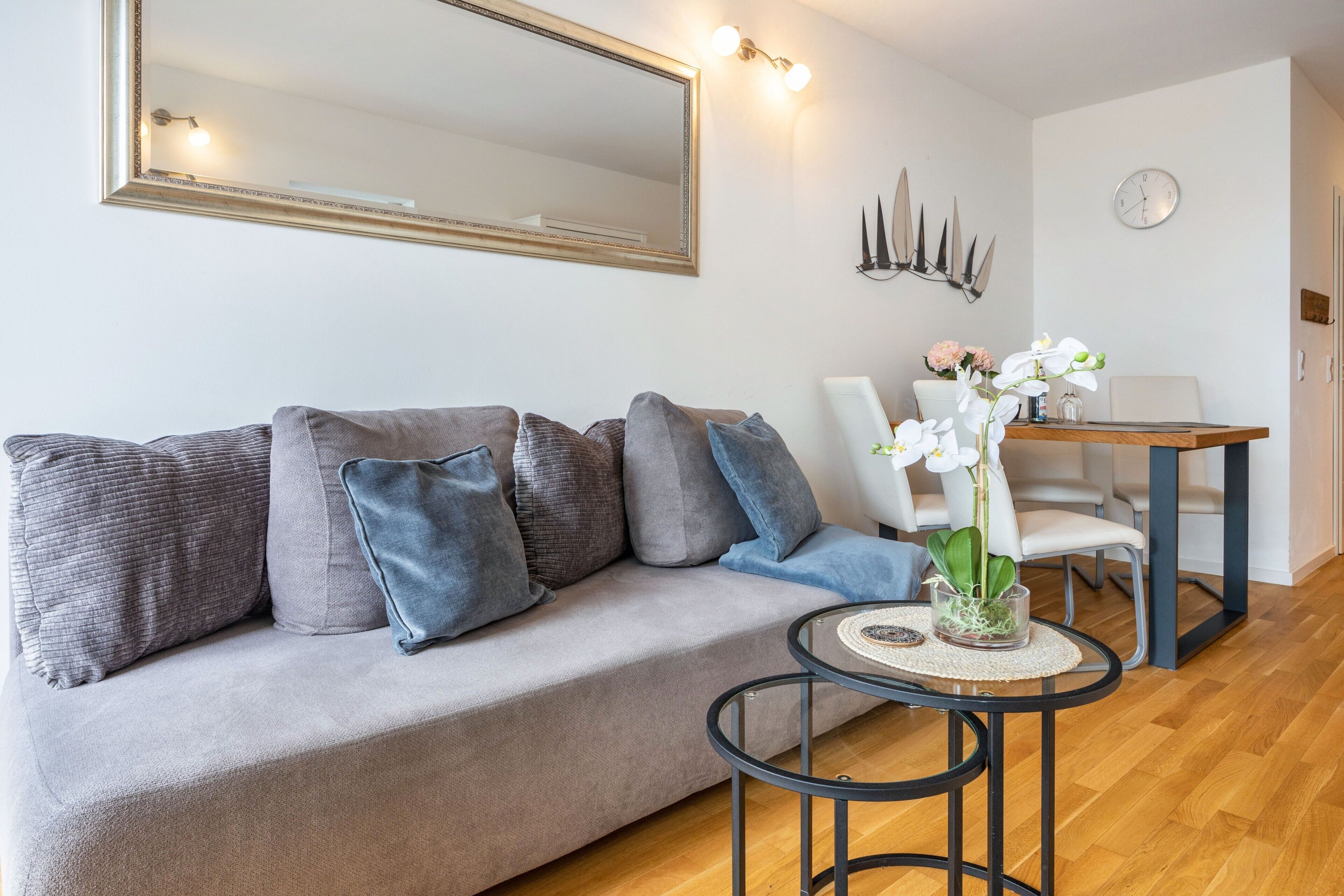 A modern living room with a grey sofa, blue cushions, a round glass coffee table, and a dining area with a wooden table and chairs.