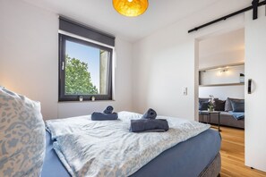 A modern bedroom with a large bed, a window with a view of trees, a small table with a plant, and a sofa in the adjacent room. - AnkerplatzSteinhuderMeer – Modern 2-Room Apartment with Balcony and Lake View (Wunstorf)