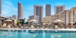 A hotel pool area with lounge chairs, umbrellas, and high-rise buildings in the background. - Waterway Balcony · 2BR Near Beach & Dining (Hallandale Beach)
