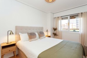 A neatly made bed with a headboard, a bedside lamp, a small bedside table, and a window with curtains. - This bright and modern apartment on Calle Julián Besteiro, in the heart of Tetuán, combines contemporary design, comfort, and functionality, creating a perfect space for extended stays in Madrid. (MADRID)