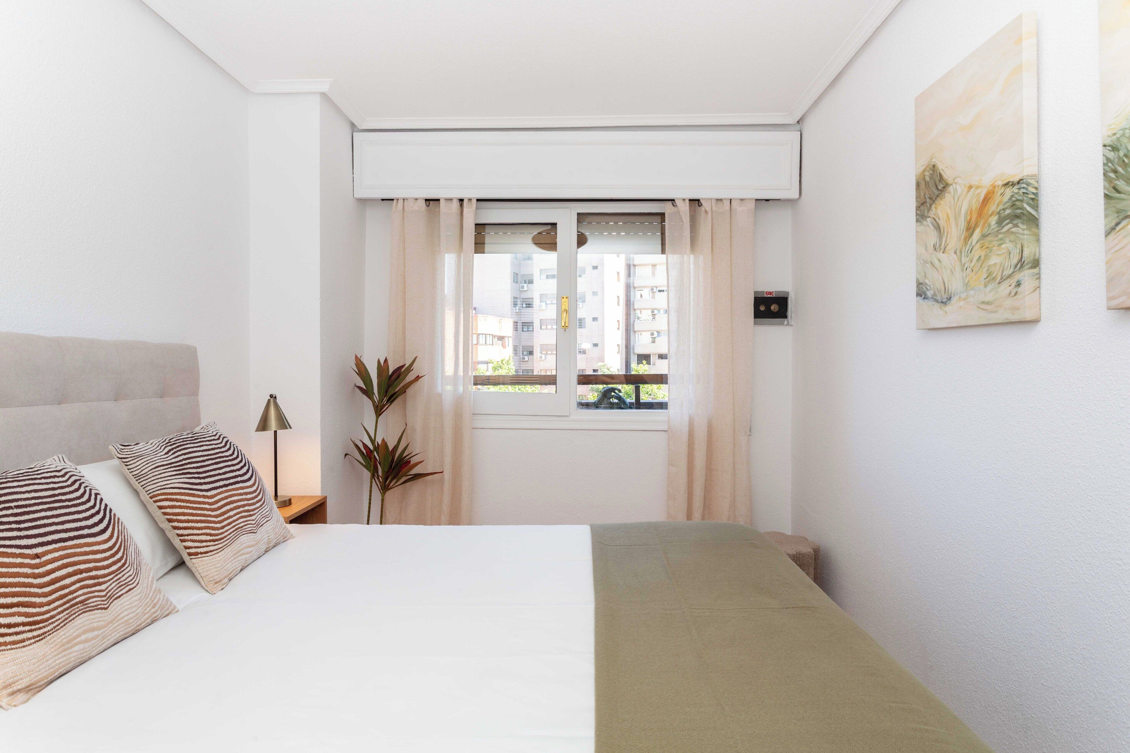 A bedroom with a bed, curtains, a window, a plant, and a painting on the wall.