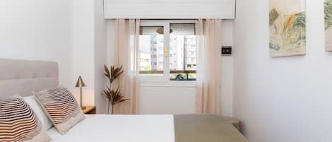 A bedroom with a bed, curtains, a window, a plant, and a painting on the wall.
