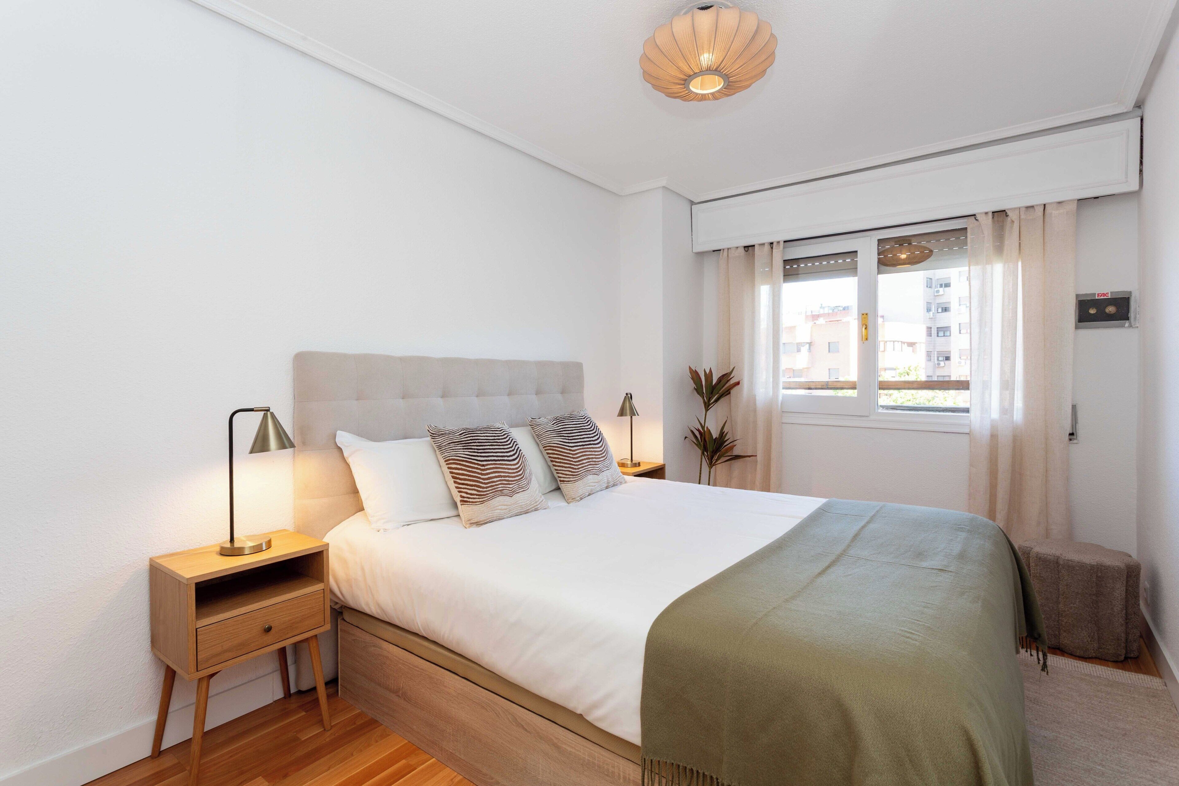 A bedroom with a bed, bedside tables, a nightstand, a window with curtains, and a ceiling light fixture.