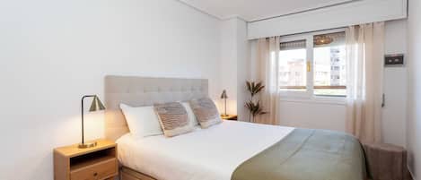 A bedroom with a bed, bedside tables, a nightstand, a window with curtains, and a ceiling light fixture.