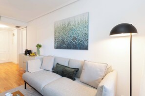 A living room with a sofa, a lamp, a painting, and a small cabinet. - This bright and modern apartment on Calle Julián Besteiro, in the heart of Tetuán, combines contemporary design, comfort, and functionality, creating a perfect space for extended stays in Madrid. (MADRID)