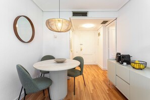 A compact room with a round table, two chairs, a hanging light fixture, a white door, and a small kitchenette area with a coffee maker and fruit bowl. - This bright and modern apartment on Calle Julián Besteiro, in the heart of Tetuán, combines contemporary design, comfort, and functionality, creating a perfect space for extended stays in Madrid. (MADRID)
