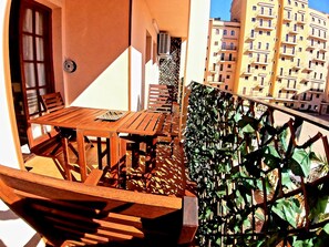 Un balcone con tavolo e sedie in legno, una pianta in vaso e vista su un edificio con diversi balconi.
