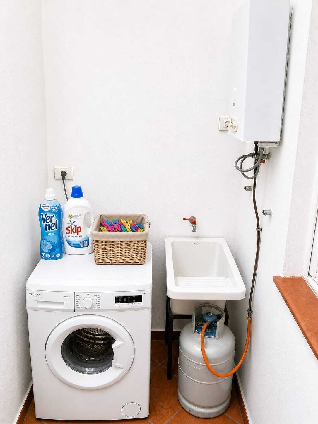 Un espace réduit dédié à la buanderie, comprenant un lave-linge, une bouteille de lessive, une bouteille d’adoucissant, un panier rempli de pinces à linge colorées, un évier et un chauffe-eau.