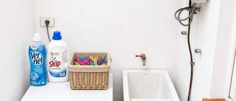 Un espace réduit dédié à la buanderie, comprenant un lave-linge, une bouteille de lessive, une bouteille d’adoucissant, un panier rempli de pinces à linge colorées, un évier et un chauffe-eau.