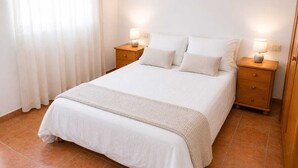Un lit bien fait, avec une couette blanche et des oreillers, deux tables de chevet avec des lampes et une fenêtre ornée de rideaux légers.