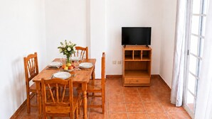 Un coin repas avec une table en bois dressée pour deux personnes, un téléviseur posé sur un meuble en bois et une fenêtre agrémentée de rideaux.