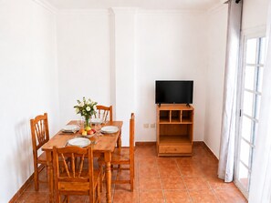 Ein Essbereich mit einem Holztisch, gedeckt für zwei Personen, einem Fernseher auf einem Holzständer und einem Fenster mit Vorhängen.