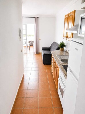 Eine kompakte Küche mit Kühlschrank, Herd und Spüle. Ein graues Sofa und ein kleiner Tisch mit einer Pflanze sind vorhanden. Eine Tür führt zu einem Außenbereich mit einem Stuhl.