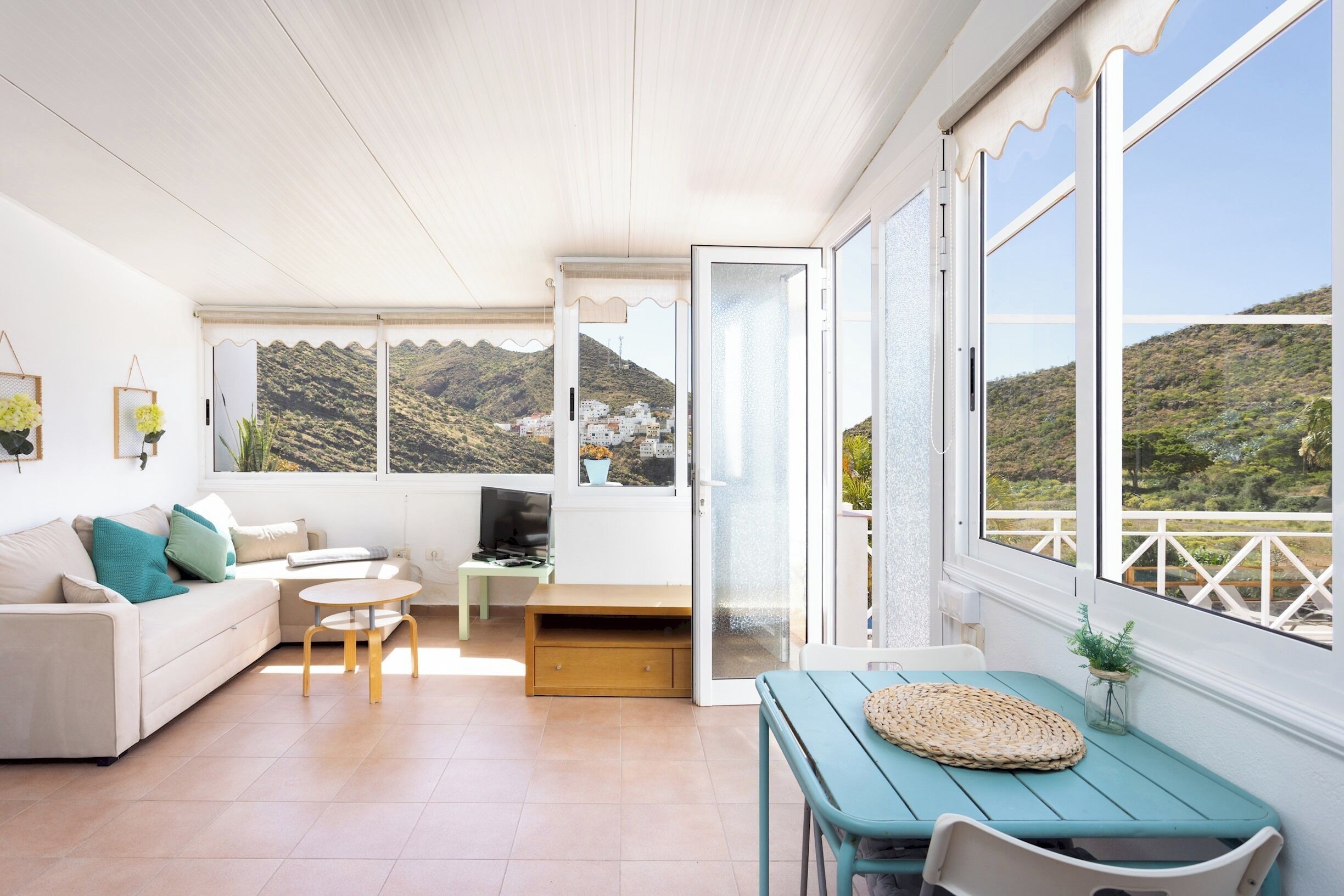 A modern living room with a sofa, a small table, a TV, and a balcony with a mountain view.