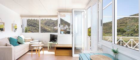 A modern living room with a sofa, a small table, a TV, and a balcony with a mountain view.