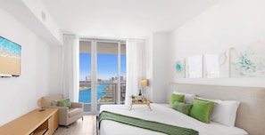 A modern hotel room with a large bed, a desk, a chair, and a view of a cityscape through large windows. - 1BR - Intracoastal View  Balcony (Hallandale Beach)