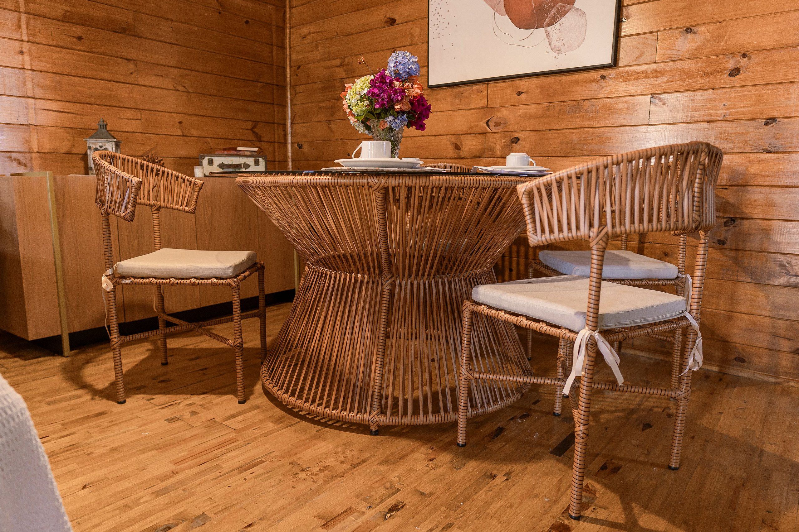 Ein Zimmer mit Holzvertäfelung, einem runden Korbstuhl, zwei Korbstühlen, einer kleinen weißen Tischlampe und einer Vase mit Blumen.