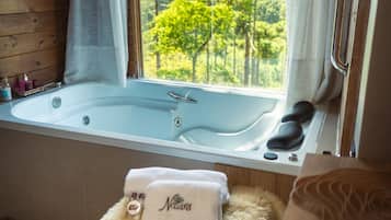 Ein Zimmer mit einer großen Badewanne, einem flauschigen weißen Hocker und Blick auf Bäume durch ein Fenster.