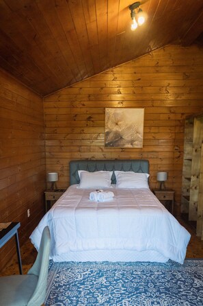 Un dormitorio con paneles de madera, una cama, dos mesitas de noche, una silla y una lámpara fijada a la pared.