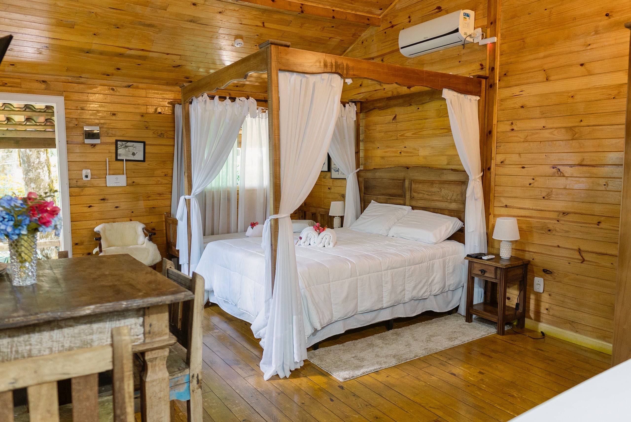 Quarto de cabana de madeira com uma cama de dossel, uma mesinha e uma cadeira.