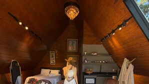 Una habitación cálida en el ático con paredes de madera, un candelabro, una mesa de comedor con sillas, un sofá y una alfombra grande y mullida.
