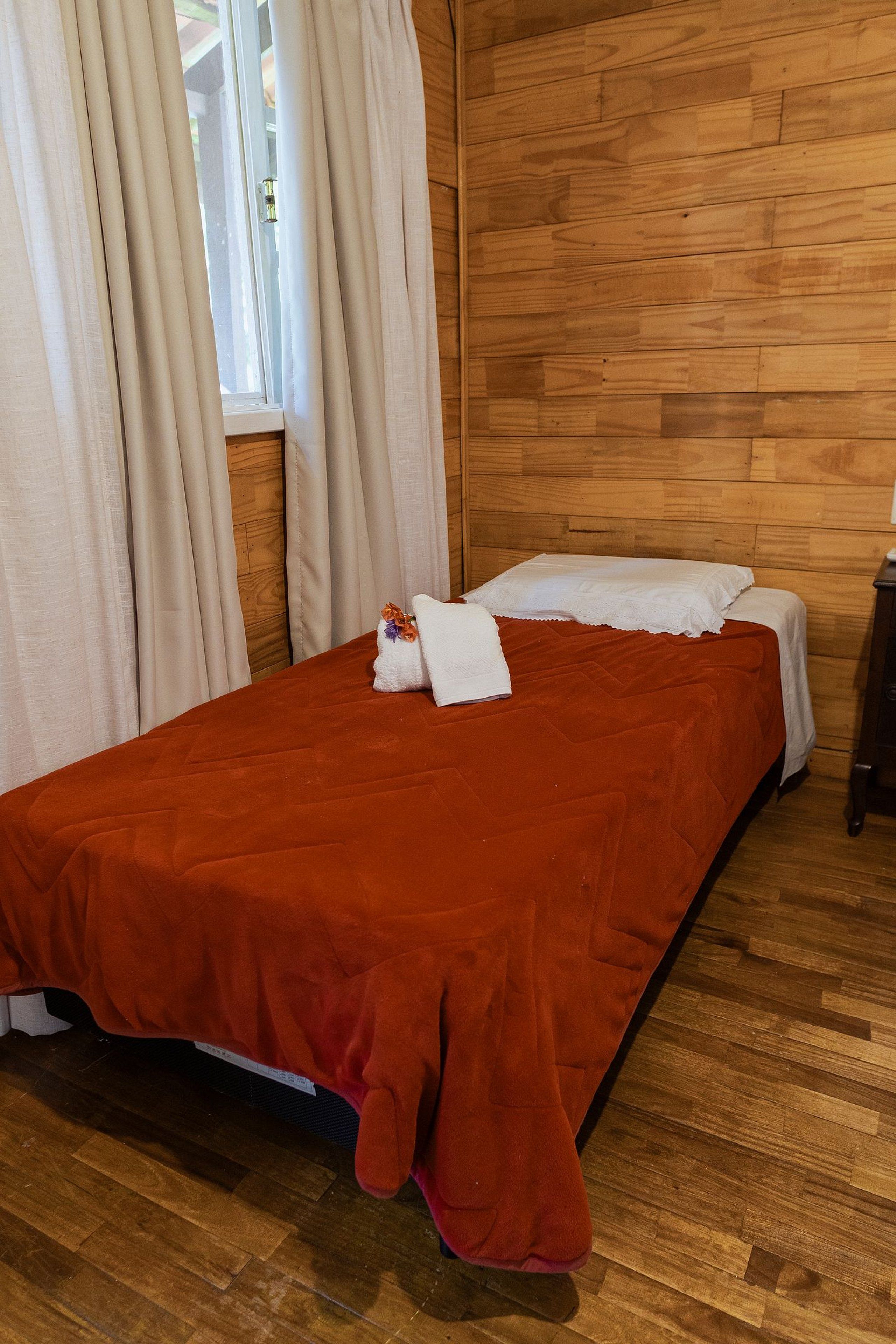 A room with a wooden wall, a bed with a red bedspread, white pillows, and a window with curtains.