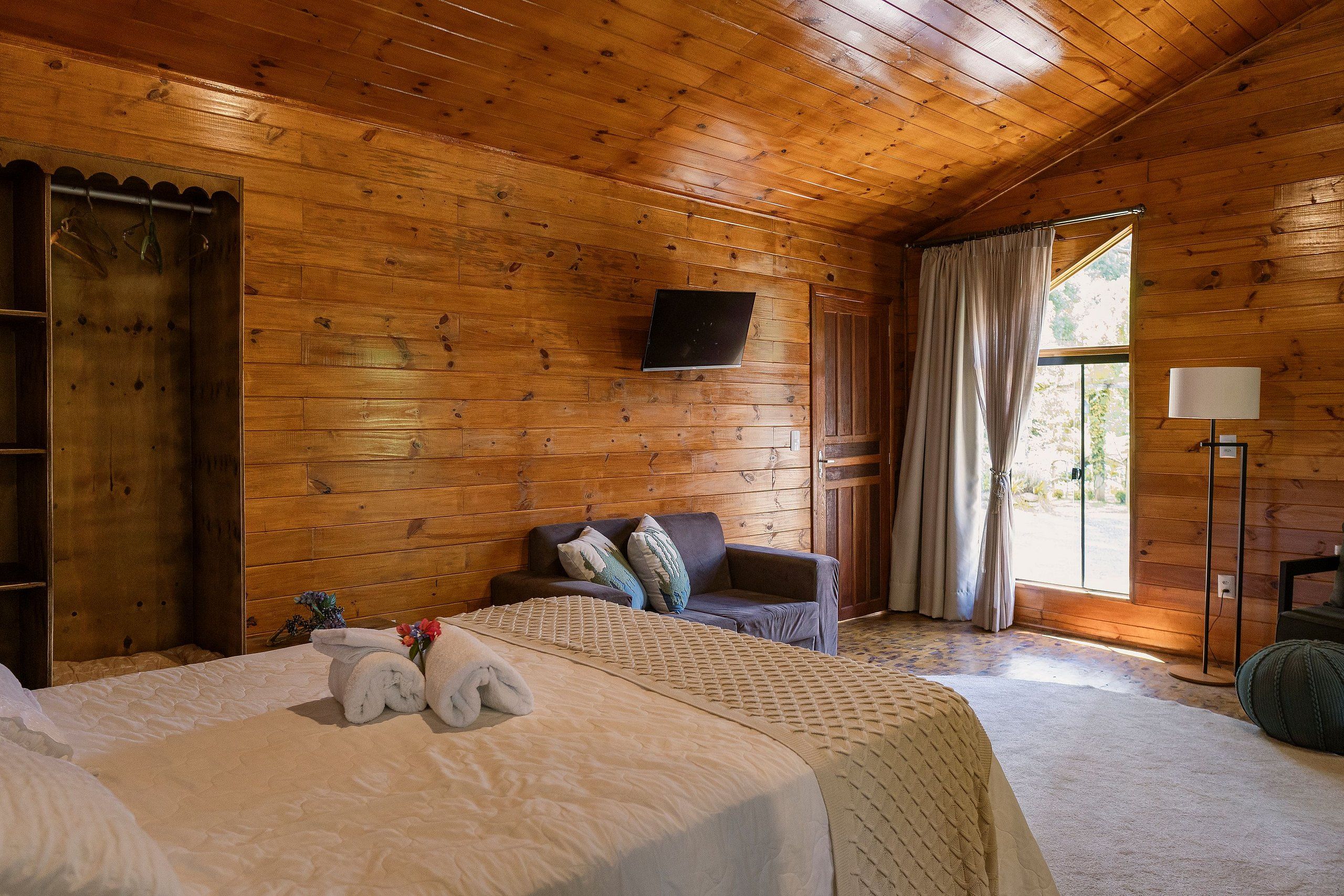 A wooden cabin room with a bed, a sofa, a TV, and a window with curtains.