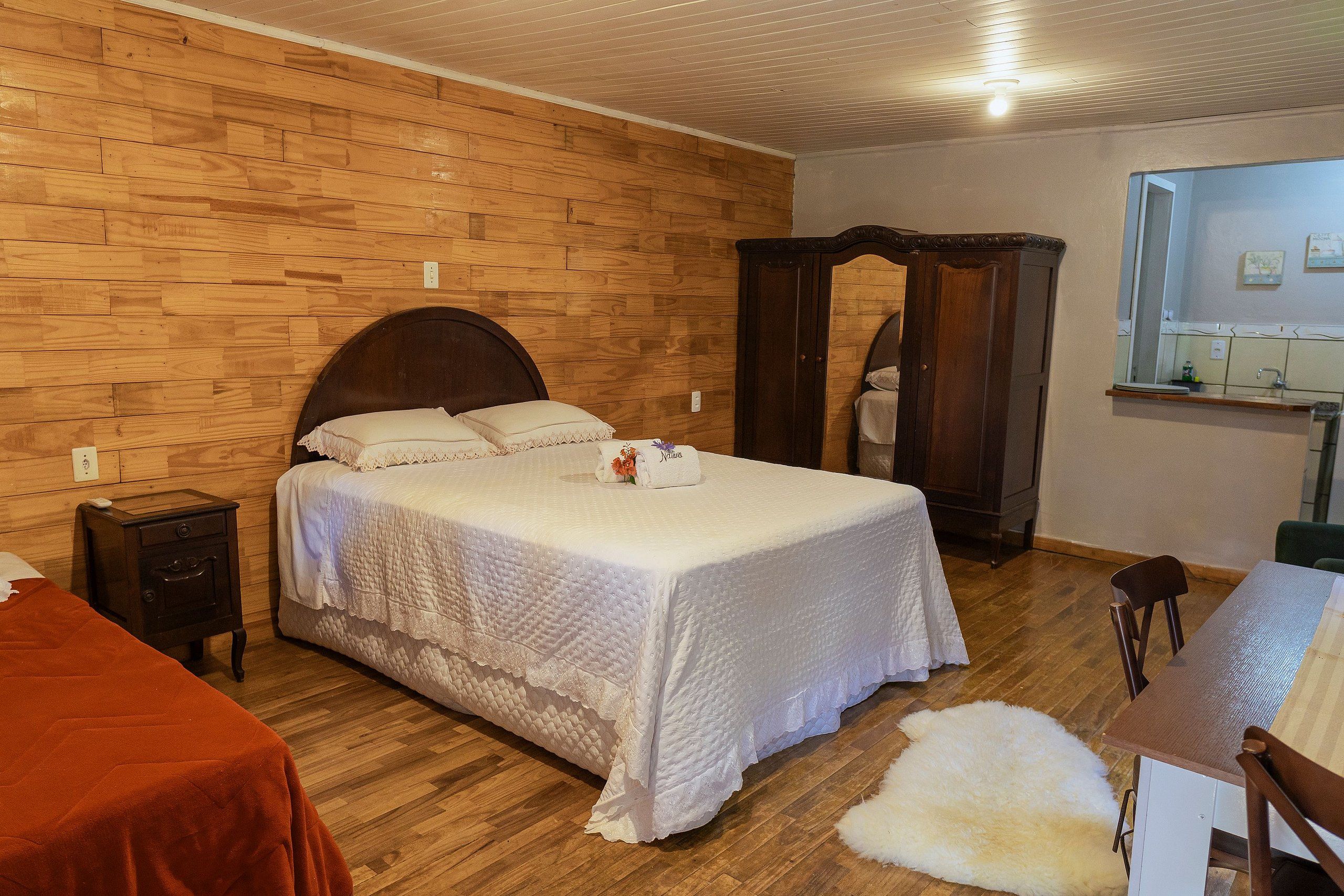 Un dormitorio con pared de madera, una cama con cubrecama blanco, un armario de madera, una mesita con una silla y un espejo sobre la mesita.