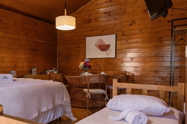 Una habitación de cabaña de madera con dos camas, una mesa de comedor, sillas de mimbre, un televisor colgante y un cuadro en la pared.