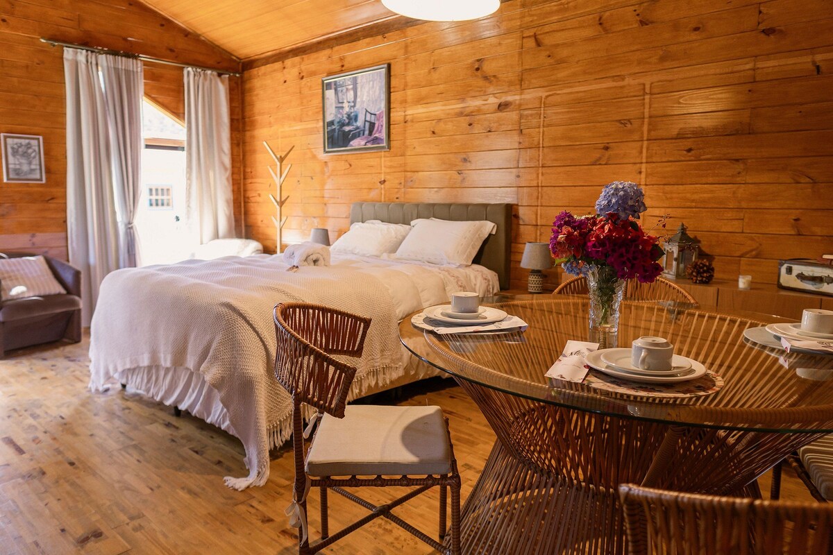 Una cabaña de madera con cama, sillas de mimbre, una mesa de cristal y un jarrón con flores.